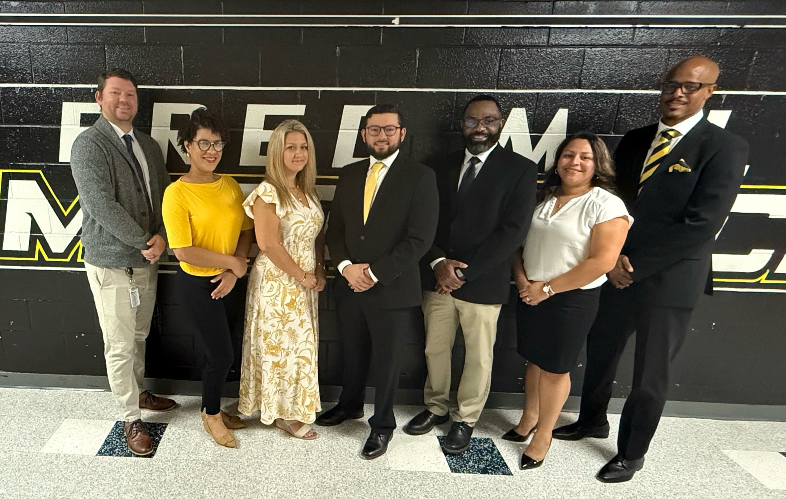 A group of photo of the admin team dressed in formal attire, standing together in front of a black and yellow backdrop, likely representing a school or organization.